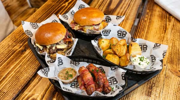 Two burgers, fried potatoes, and bacon-wrapped sausages with dipping sauces on a wooden table.