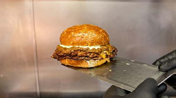 Double smashburger with cheese, sauce, and a sesame seed bun on a metal spatula.