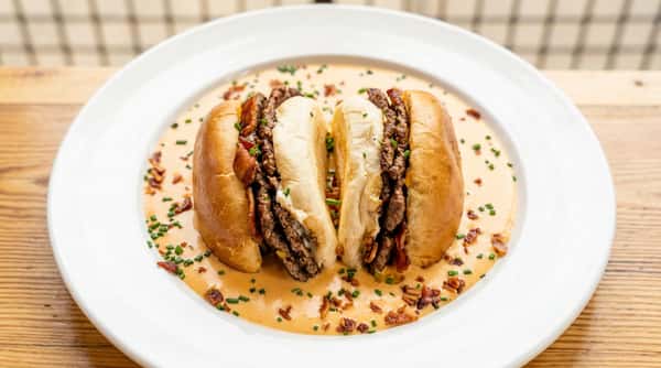 Cheesesteak sandwich submerged in creamy orange sauce, topped with bacon bits and chives on a white plate.