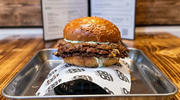 Double smash burger on a sesame bun with sauce, sitting on branded paper in a metal tray. Menu visible behind it.