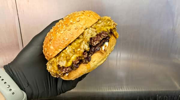 A hand in a black glove holds a sesame seed bun burger with two dark brown patties, melted yellow cheese, and green relish.