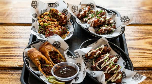 Four baskets of chicken wings and egg rolls on a black tray, arranged on a wooden surface.