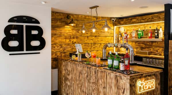 A modern bar interior with a distressed wood counter and back wall, beer taps, liquor bottles on shelves, and a neon "Bar" sign.