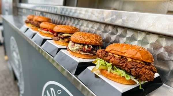 A row of four sandwiches: a patty melt with bacon and melted cheese, and a crispy fried chicken sandwich with lettuce and pickles.