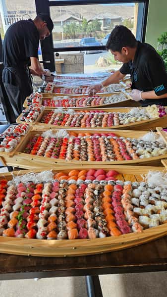 Two sushi chefs arranging a large assortment of colorful sushi rolls on wooden boat displays.