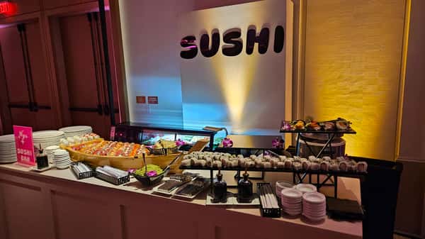 Sushi buffet with assorted rolls, serving dishes, and a large "SUSHI" sign in the background.