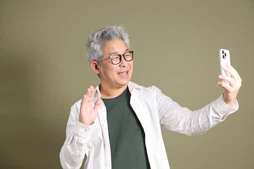 A smiling middle-aged man with gray hair and glasses is holding up a smartphone and waving, appearing to be on a video call. He is wearing a white shirt over a dark green t-shirt, standing against a plain light olive background.