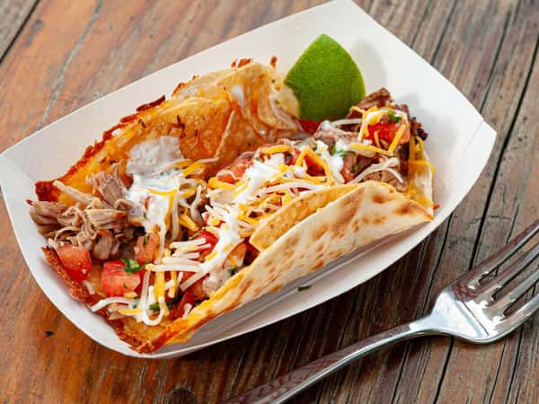 Two crispy grilled tacos filled with seasoned shredded meat, pico de gallo, melted cheese, and creamy sauce, served with a fresh lime wedge at Burrito Famous.