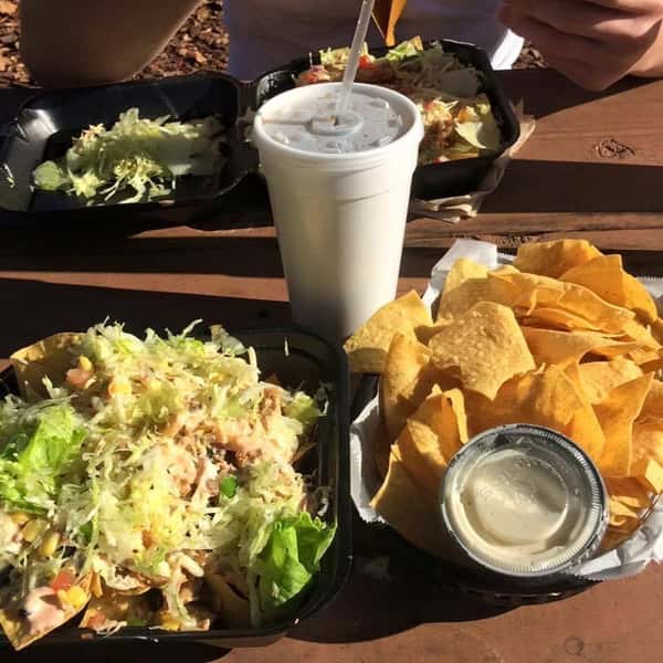 Outdoor meal at Burrito Famous featuring loaded fries, a fresh salad, crispy tortilla chips with queso, and a fountain drink on a sunny patio table.