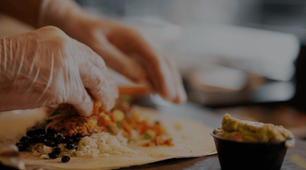 Hands wearing food-safe gloves assembling a fresh Burrito Famous burrito with seasoned meat, rice, black beans, salsa, and guacamole on a tortilla in the restaurant kitchen.