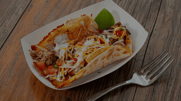 Two crispy grilled tacos filled with seasoned shredded meat, pico de gallo, melted cheese, and creamy sauce, served with a fresh lime wedge at Burrito Famous.