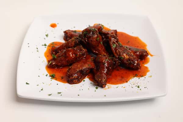 This image displays a restaurant meal. Crispy chicken wings are smothered in a vibrant red sauce and garnished with fresh parsley, and served on a square plate.