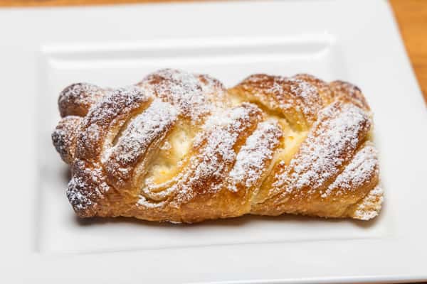 Close-up of a freshly baked golden-brown pastry topped with powdered sugar and drizzled icing, served on a white plate at a café.