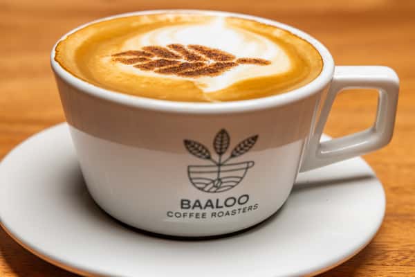 Freshly prepared cappuccino with creamy milk foam and decorative latte art, served in a branded white ceramic coffee cup on a wooden café table.