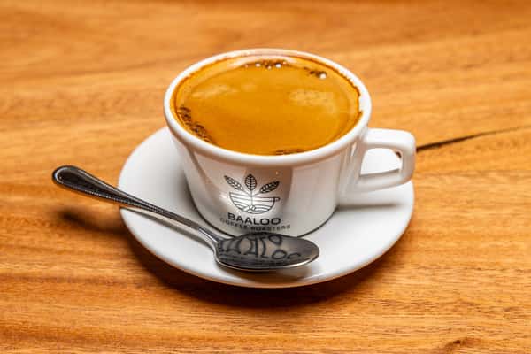 Freshly brewed espresso with a rich golden crema, served in a white ceramic cup with a spoon on a saucer atop a warm wooden café table.