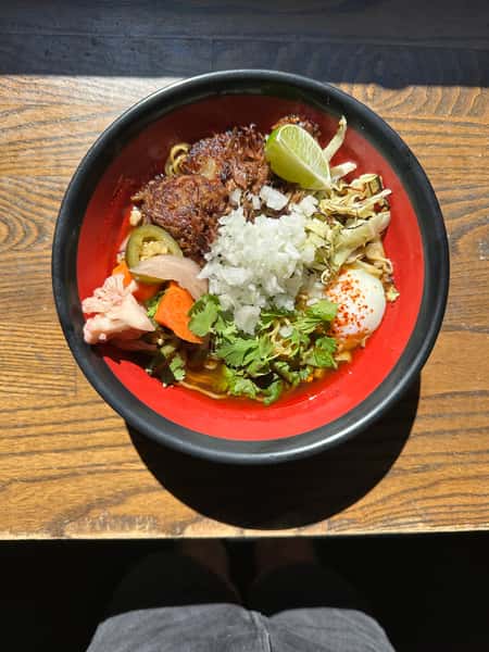 Bowl of ramen with shredded meat, soft-boiled egg, onions, lime, cilantro, and pickled vegetables