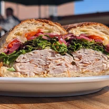 Turkey sandwich with lettuce, pickled onions, and roasted red peppers on crusty bread, served on a white plate outdoors.