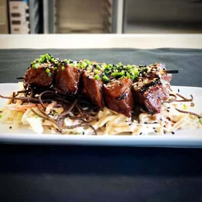 Grilled beef skewers topped with chopped chives, served over a bed of crispy noodles and creamy coleslaw on a white plate.