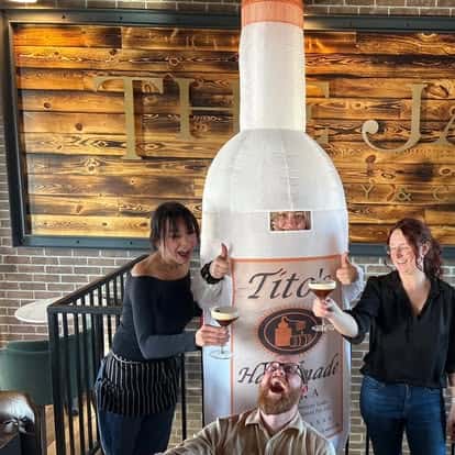 Three people posing with drinks, one dressed in an inflatable Tito's Handmade Vodka bottle costume, inside The James bar.