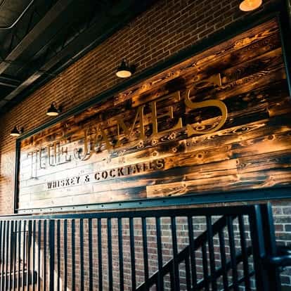 The image shows a rustic wooden wall sign for "The James" with the text "Whiskey & Cocktails" beneath it. The sign is mounted on a brick wall and illuminated by multiple overhead lights, giving it a warm, inviting atmosphere. There's a black metal railing in front, suggesting this is part of an entrance or patio area for the establishment.