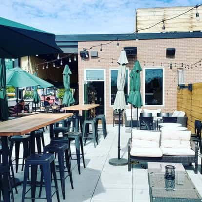 Sunny outdoor patio with high tables, bar stools, cushioned seating, and green umbrellas under string lights.