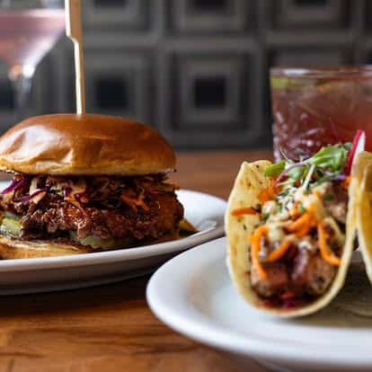 Burger with coleslaw and pickles alongside a plate of tacos filled with grilled meat, vegetables, and fresh greens.
