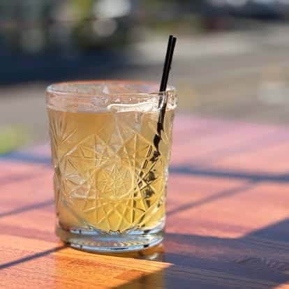 Golden cocktail served over ice in a textured glass with a black straw, placed on a sunlit wooden table.