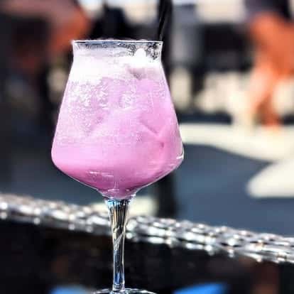 Vibrant lavender cocktail served in a stemmed glass with ice, set against a blurred outdoor background.