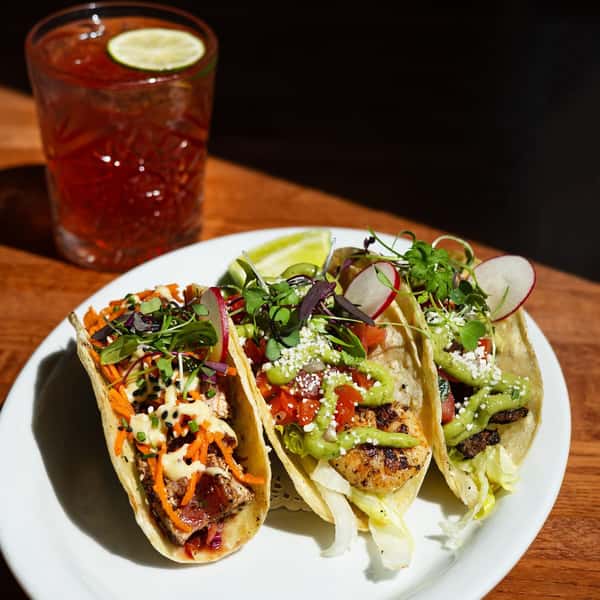 Trio of gourmet tacos with fresh vegetables, grilled seafood, and vibrant garnishes, served with a lime wedge and cocktail.