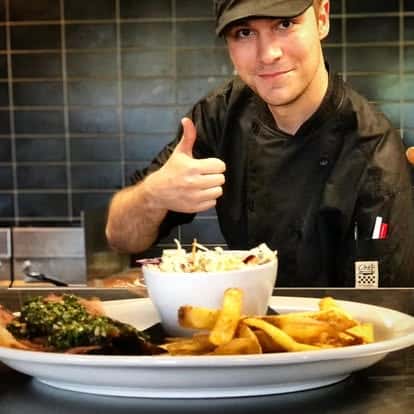 Smiling chef in black uniform giving a thumbs-up behind a plate of fries, coleslaw, and grilled meat topped with herbs.