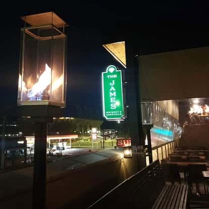 Night view of The James rooftop patio with fire lamps, outdoor seating, and a glowing green sign overlooking the street.