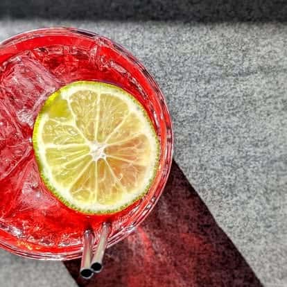 Vibrant red cocktail with ice, garnished with a fresh lime slice, served with two straws on a sunlit surface.