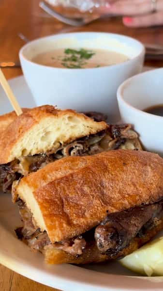 Toasted baguette sandwich filled with sliced beef and onions, served with au jus and a creamy soup garnished with herbs.