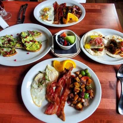 Assorted breakfast plates with bacon, eggs, avocado toast, breakfast tacos, roasted potatoes, and fresh fruit on a wooden table.