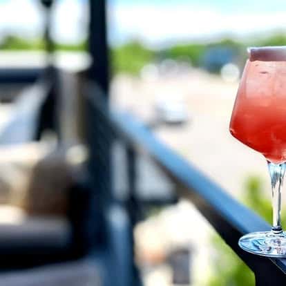 Pink cocktail in a stemmed glass on an outdoor patio railing with a blurred city street in the background.