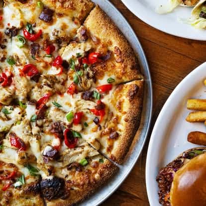 Close-up of a freshly baked pizza topped with chicken, red peppers, and green onions, with a burger and fries on the side.