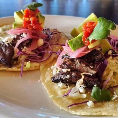 Two soft tacos filled with grilled steak, shredded cabbage, pickled onions, avocado chunks, crumbled cheese, a dollop of red salsa, and fresh cilantro.