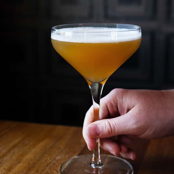 A hand holding a coupe glass filled with a golden-brown cocktail topped with a thin layer of foam.