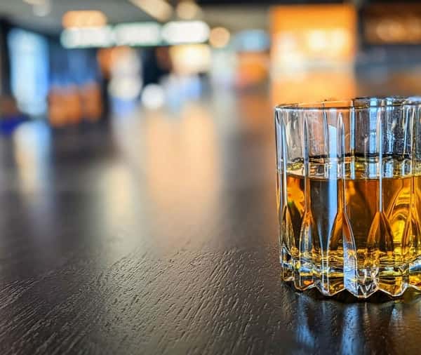 A glass of amber whiskey on a dark wooden bar with a softly blurred background of warm lights.