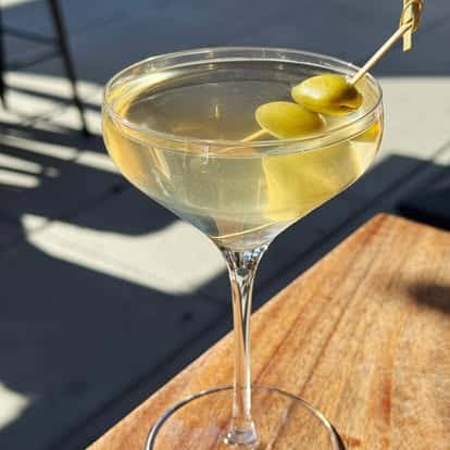 Classic martini garnished with three green olives on a cocktail pick, served in a coupe glass on a sunny outdoor table.