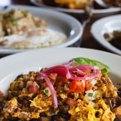 Scrambled eggs with diced vegetables, pickled onions, and avocado slices served on a white plate with breakfast dishes in the background.