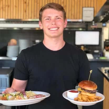 The image shows a smiling person wearing a black shirt, holding two plates — one with tacos and the other with a burger, onion rings, and a side of dipping sauce. The background suggests a restaurant setting.