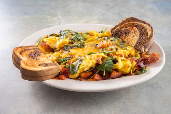 Scrambled eggs with spinach, cheese, tomatoes, and bacon served with toasted marble rye bread on a white oval plate.