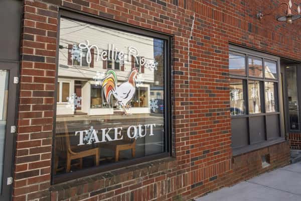 Brick storefront window with Nellie Rose Take Out lettering and rooster graphic, reflecting a small-town street scene.