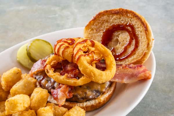 Cheeseburger with bacon, crispy onion rings, and barbecue sauce, served with tater tots and pickle slices on a white plate.