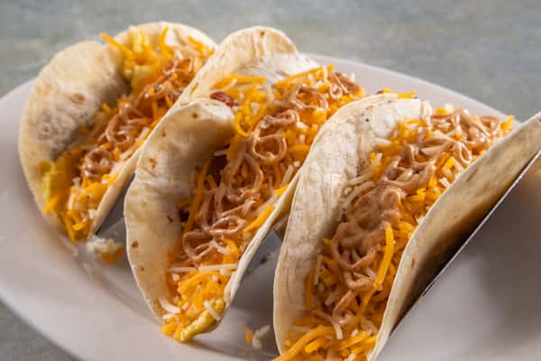 These appear to be breakfast tacos made with soft flour tortillas, scrambled eggs, shredded cheddar and Monterey Jack cheese, bacon or sausage, and a drizzle of creamy sauce—likely chipotle or spicy mayo. A flavorful and hearty way to start the day!