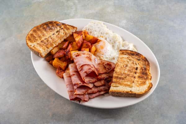 Breakfast plate with grilled ham, over-easy eggs, home fries, and toasted bread on a white oval plate.