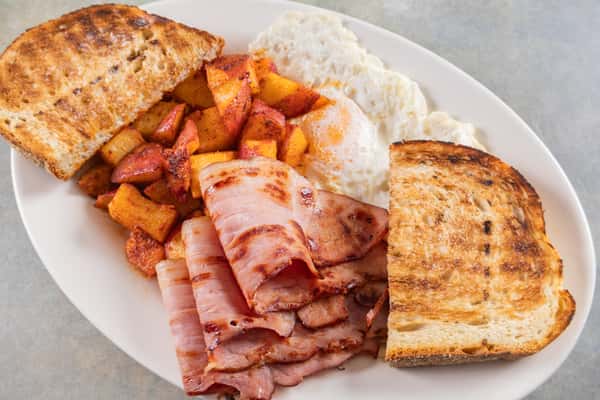 Classic breakfast plate with grilled ham slices, home-fried potatoes, two sunny-side-up eggs, and toasted sourdough bread.