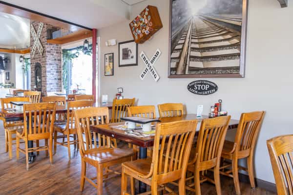 Cozy diner interior with wooden tables and chairs, decorated with railroad signs, framed photos, and a dramatic train track painting.