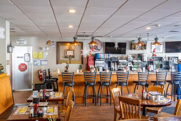 A cozy diner interior with wooden furniture, a long bar counter lined with high chairs, and festive winter decorations like snowflakes and small Christmas trees. The space includes a coffee station and multiple flat-screen TVs mounted on the wall.
